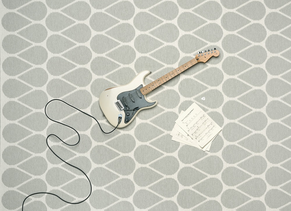 Auf dem Foto sieht man den Teppich Otis Linen von Pappelina. Auf dem Kunststoffteppich liegt eine E-Gitarre und daneben Notenblätter und ein schwarzes Kabel schlängelt sich von der E-Gitarre über den Teppich.