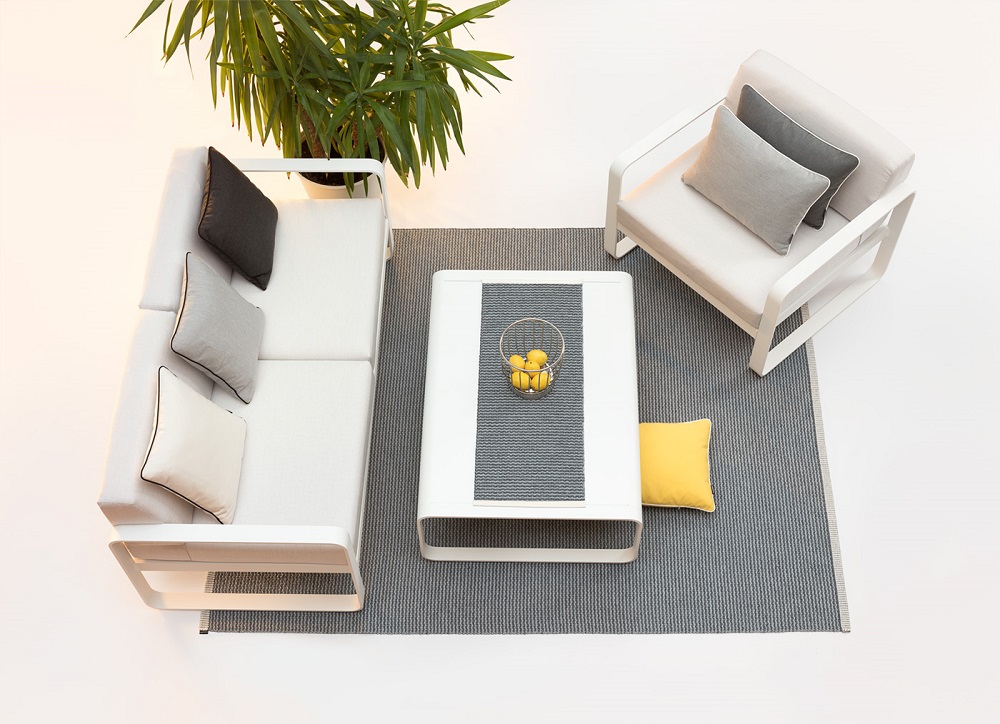 Ein grauer Pappelina-Teppich liegt auf einem hellen Untergrund. Darauf ist ein weißer Tisch, ein weißes Sofa und ein Sessel arrangiert. Das Sofa und der Sessel sind mit farblich abgestimmten Kissen dekoriert. Auf dem Tisch liegt ein grauer Pappelina Tischläufer, auf dem ein Korb mit Obst steht. Neben dem Teppich steht eine Grünpflanze.