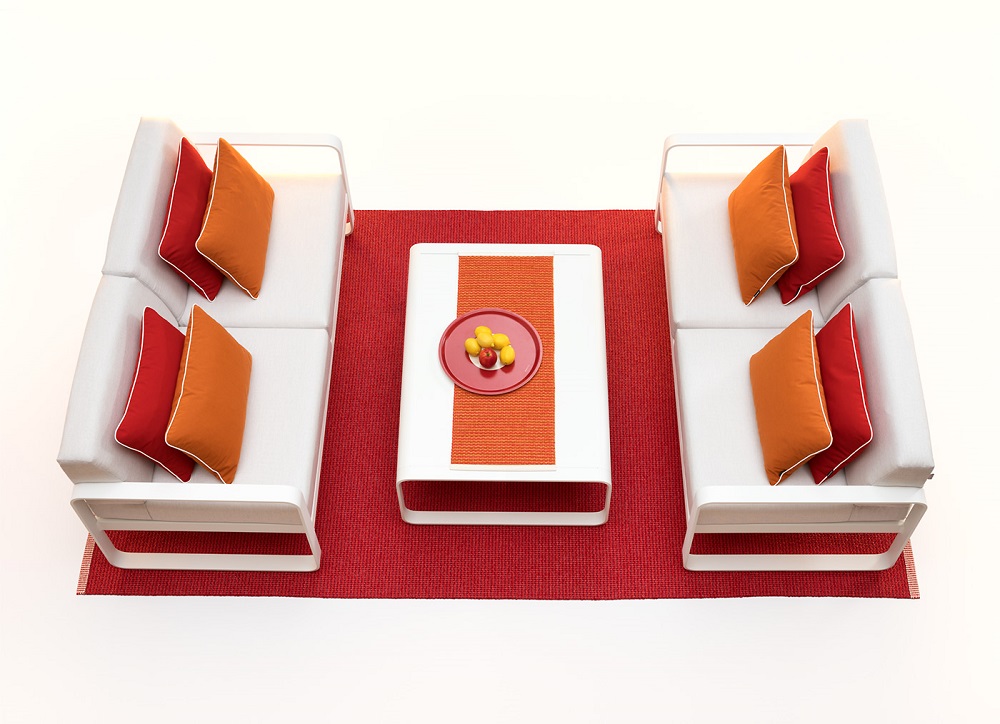 Ein roter Pappelina-Teppich liegt auf einem hellen Untergrund. Darauf ist ein weißer Tisch, ein weißes Sofa und ein Sessel arrangiert. Das Sofa und der Sessel sind mit farblich abgestimmten Kissen dekoriert. Auf dem Tisch liegt ein roter Pappelina Tischläufer auf dem eine Schale mit Obst steht.
